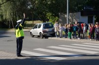 Zdjęcie kolorowe, przedstawiające policjanta ruchu drogowego pokazujące prawidłowe korzystanie z przejscia dla pieszych oraz dzieci z klasy pierwszej.