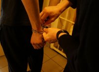 Zdjęcie kolorowe, przedstawiające policjanta, który zakłada kajdanki osobie zatrzymanej.
