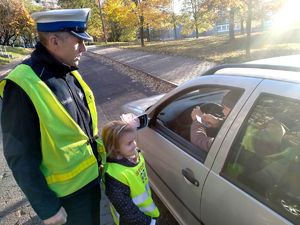 Zdjęcie przedstawiające policjanta ruchu drogowego oraz przedszkolaka stojących przy pojeździe, w trakcie sprawdzania czy pasażer w pojeździe ma zapięte pasy bezpieczeństwa.